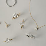 A luxurious flat-lay arrangement of KARAAT fine jewelry on a neutral grey background, featuring a rose gold emerald-cut engagement ring, an oval solitaire, a round diamond ring, a tennis bracelet, and a delicate diamond pendant necklace.