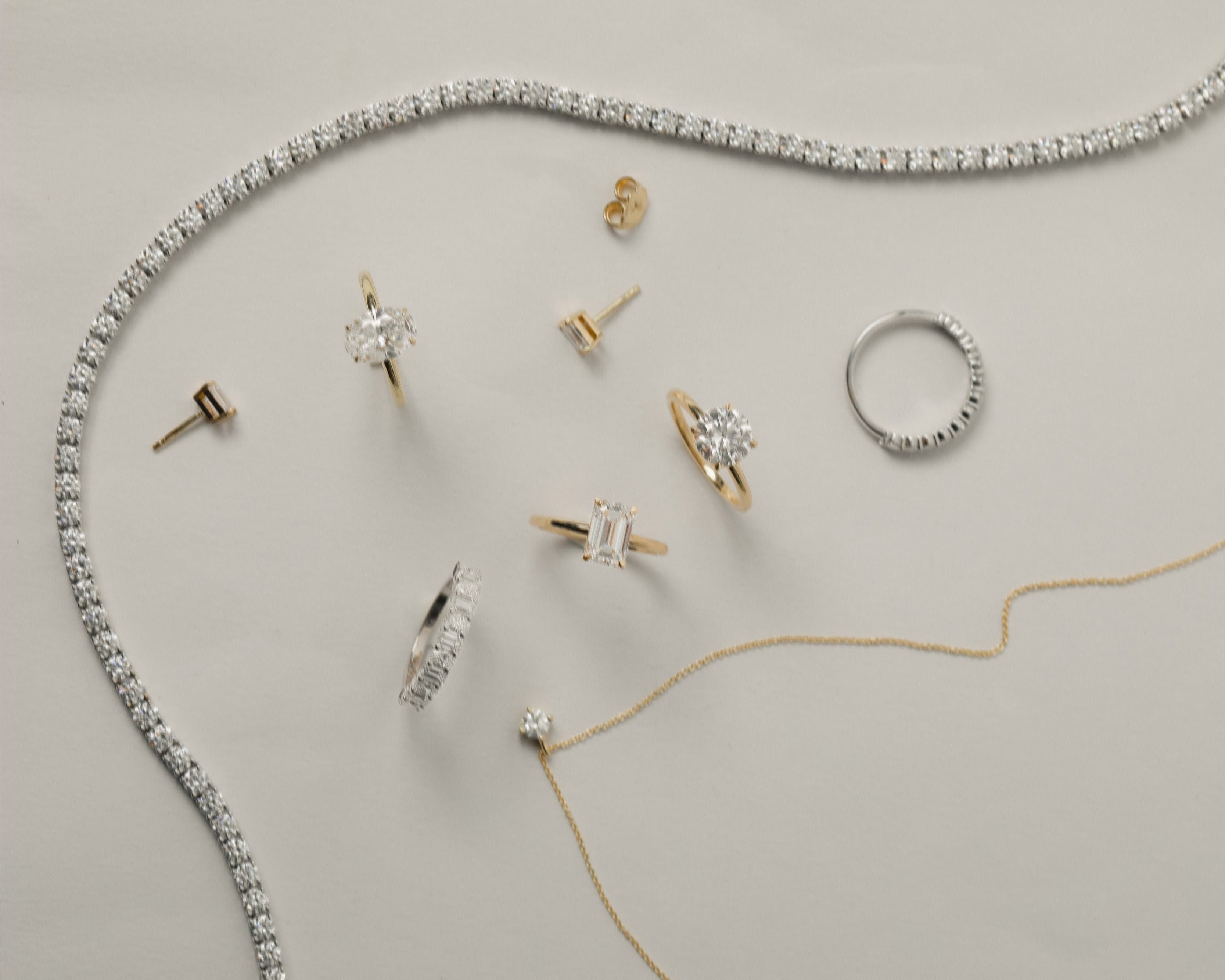 A styled flat-lay of multiple jewelry pieces on a light warm-gray background, including a white gold tennis necklace, a yellow gold pendant necklace, several rings, stud earrings, and a small pendant.
