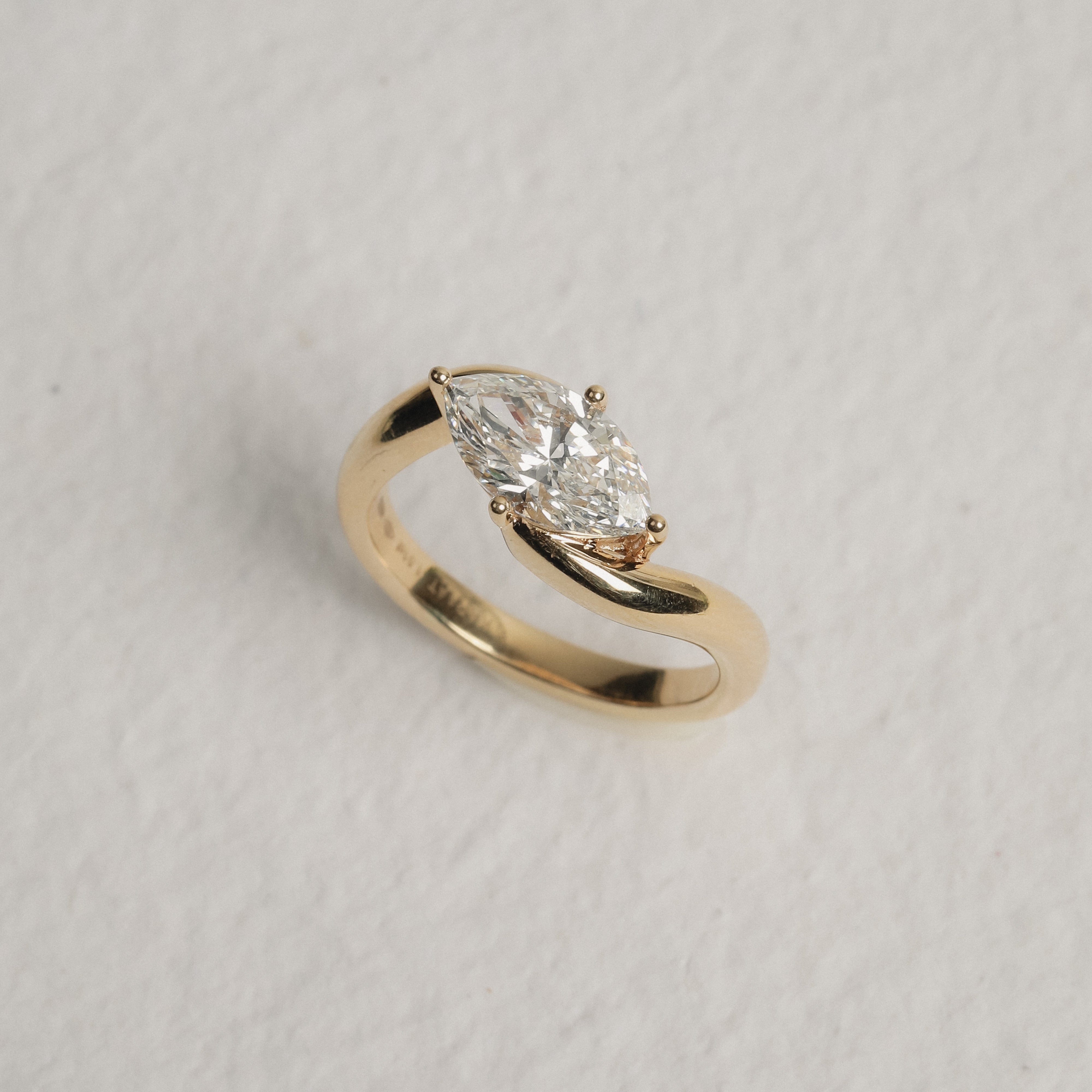 A yellow gold ring featuring a marquise-cut clear gemstone in a prong setting, displayed on a textured off-white background.