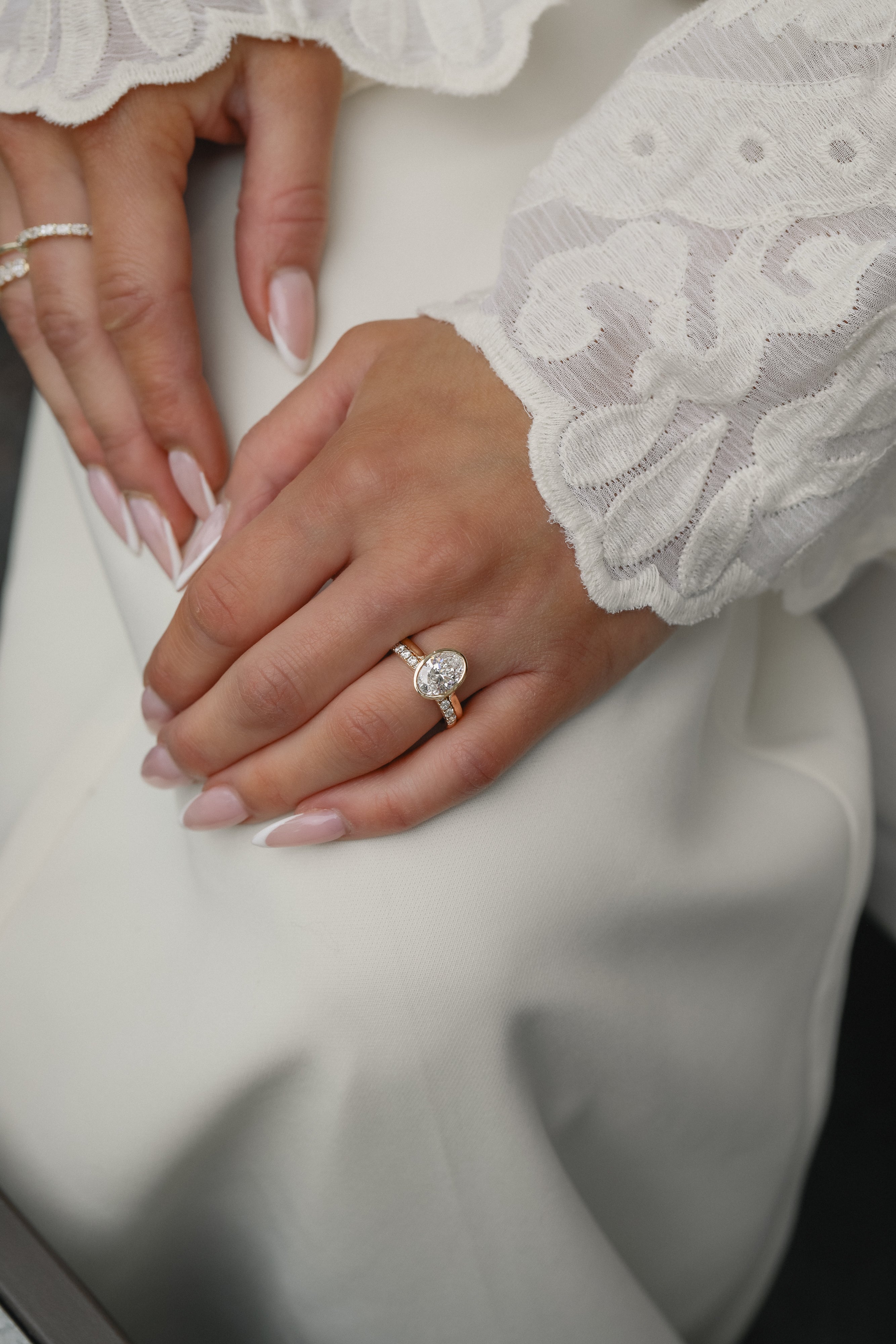 Elegant bridal lifestyle shot of a bride in a lace gown showcasing a modern bezel-set oval diamond engagement ring stacked with the Pavé Diamond Ring II in yellow gold.