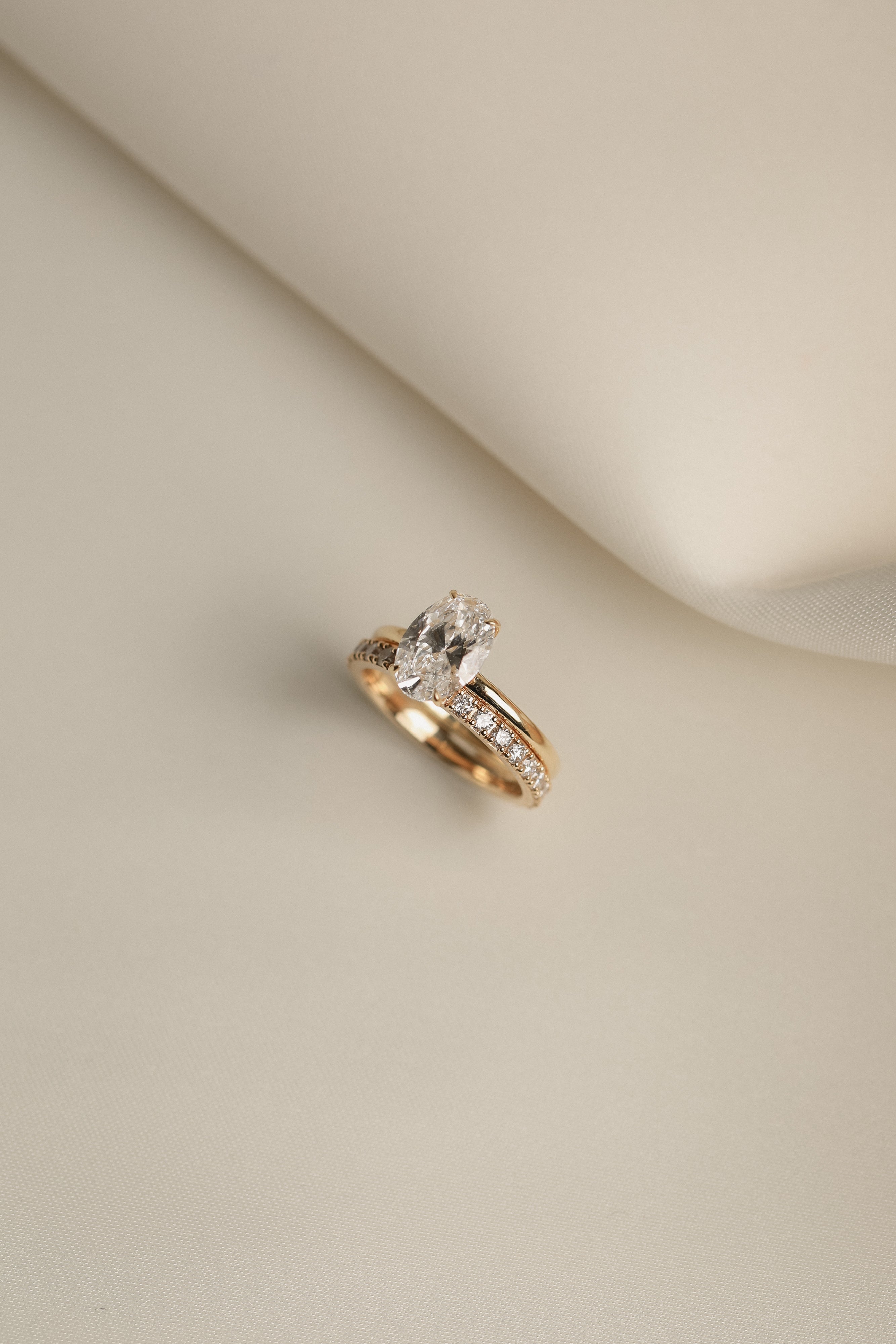 Still life shot of a luxury bridal ring stack featuring a classic oval solitaire diamond engagement ring paired flush with the Pavé Diamond Ring II on a cream fabric background.