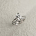 A professional still-life photograph of a KARAAT's oval-cut diamond solitaire engagement ring with a sparkling pavé band in 18k yellow gold, displayed on a neutral background.