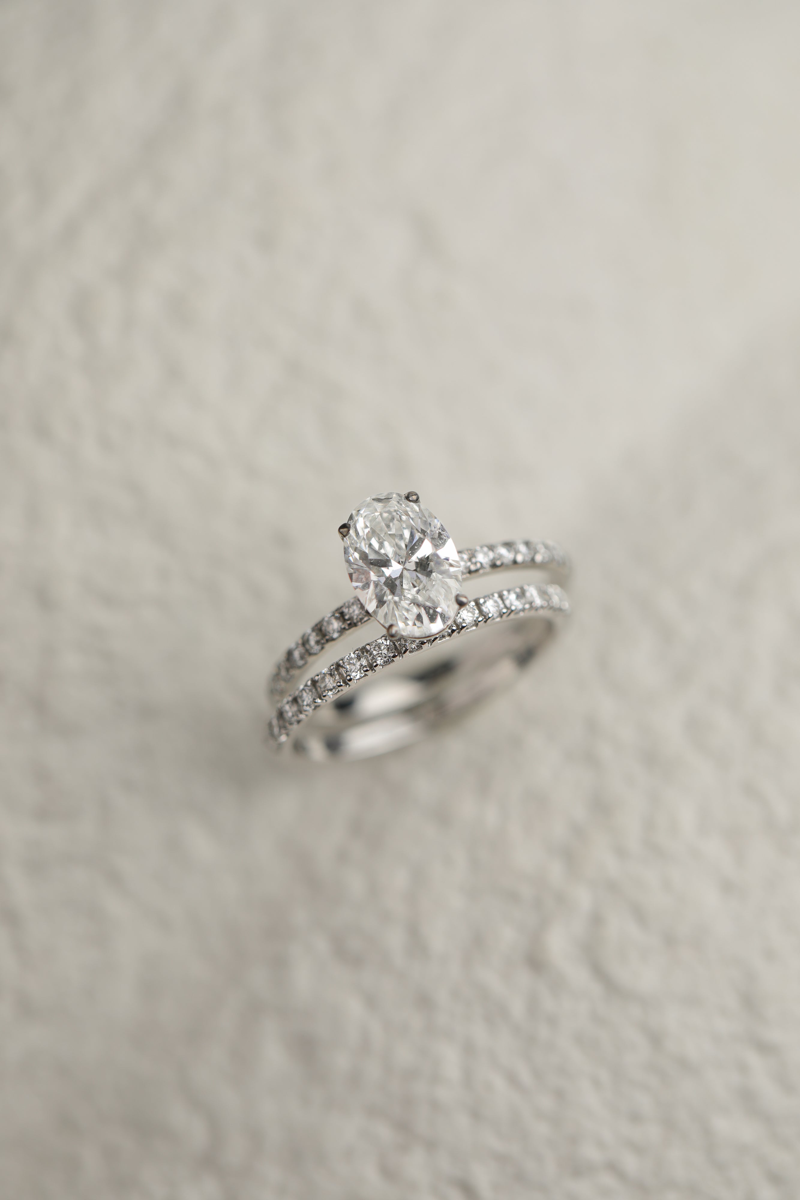 A professional still-life photograph of a KARAAT's oval-cut diamond solitaire engagement ring with a sparkling pavé band in 18k rose gold, displayed on a neutral background.