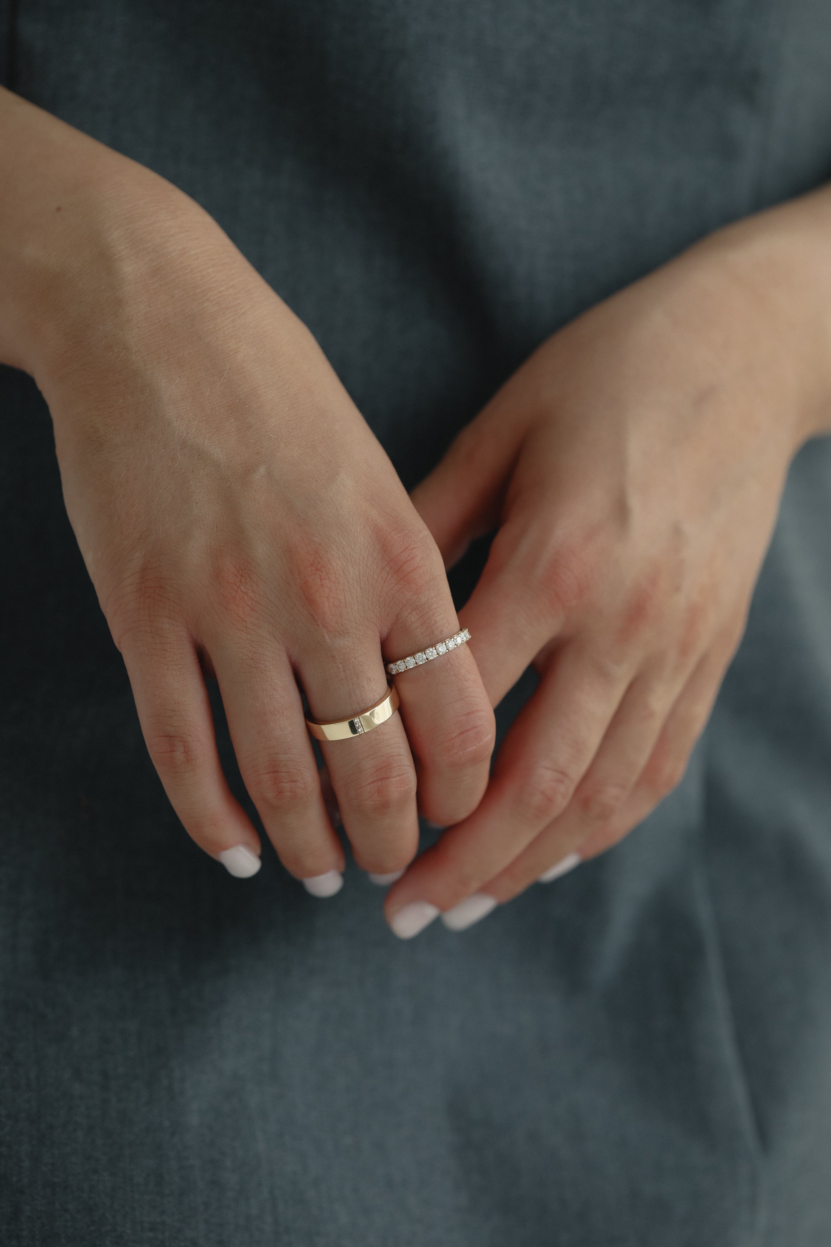 Detail shot of a model's hands gently clasped over a slate blue dress, showing a modern mixed stack featuring a wide solid gold band paired with the striking Pavé Diamond Ring III in white gold.