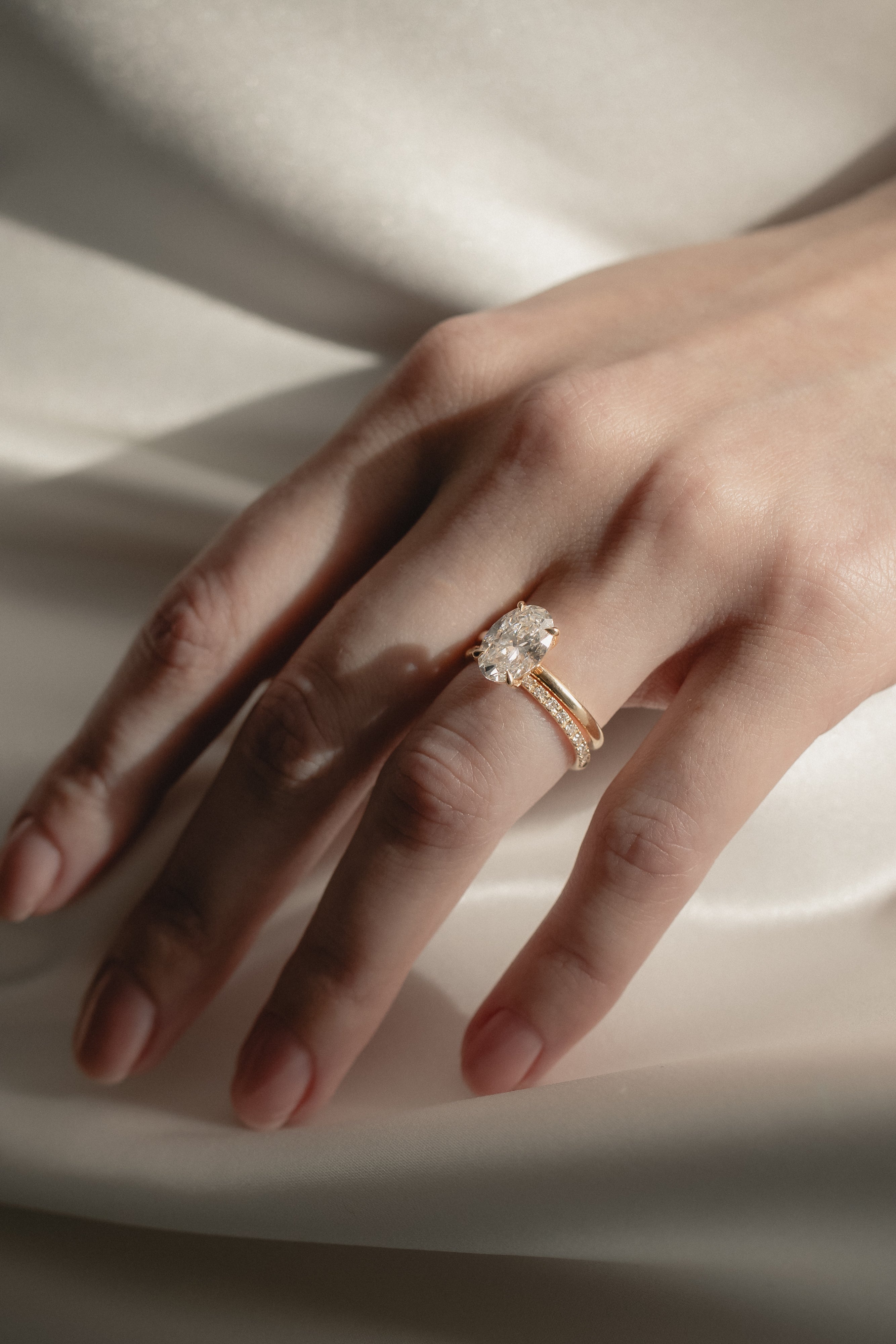 A soft-focus bridal image of a model's hand on shimmering white silk, showcasing a classic oval solitaire diamond engagement ring nested flush beside a delicate KARAAT pavé diamond wedding band.