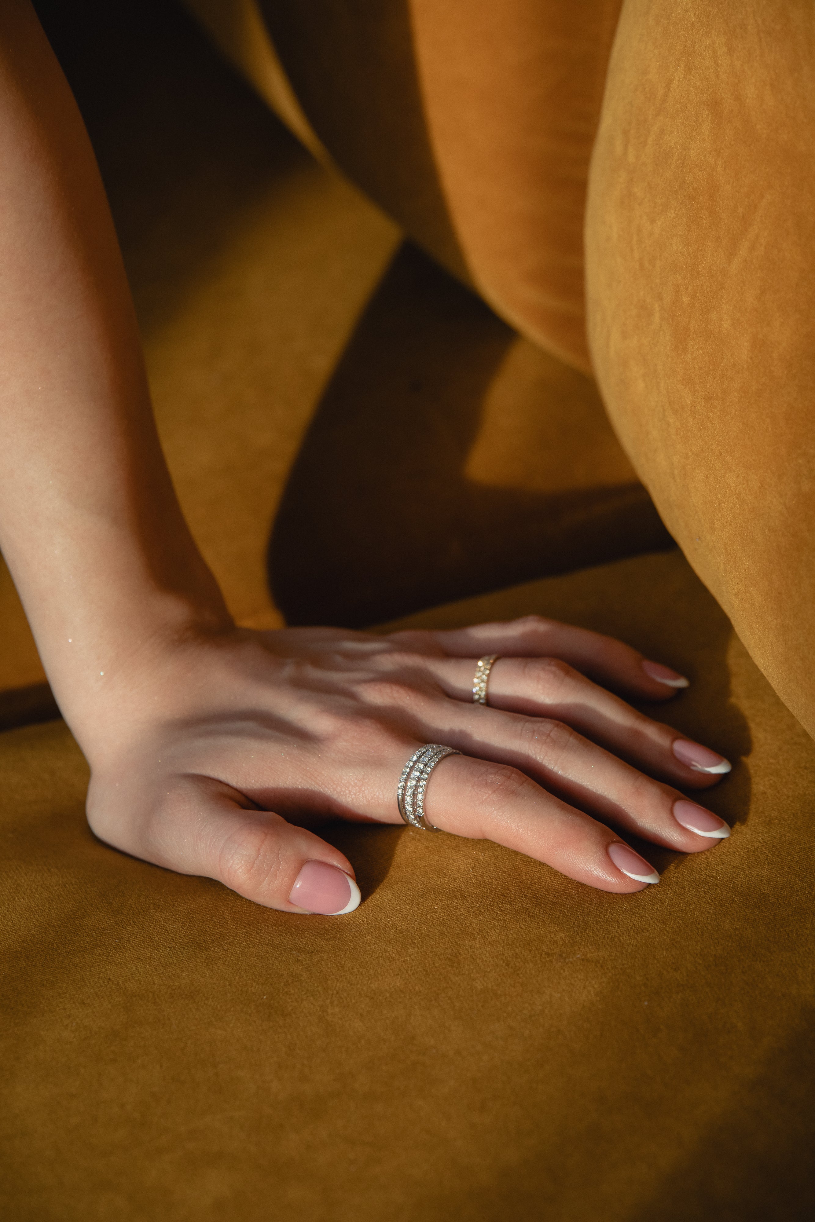 Sunlit detail shot of a model's hand resting on golden ochre fabric, showcasing a prominent ring stack featuring a thick diamond eternity band layered with a delicate classic gold pavé diamond ring.