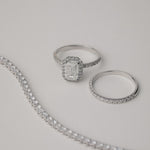 A luxury still-life photograph featuring an emerald-cut diamond halo engagement ring paired with a matching KARAAT white gold pavé diamond ring and a partial tennis bracelet on a neutral grey background.