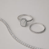 A luxury still-life photograph featuring an emerald-cut diamond halo engagement ring paired with a matching KARAAT white gold pavé diamond ring and a partial tennis bracelet on a neutral grey background.