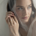 An editorial portrait of a bride looking through a sheer wedding veil, elegantly showcasing a KARAAT pear-shaped diamond solitaire engagement ring stacked with a matching rose gold wedding band.