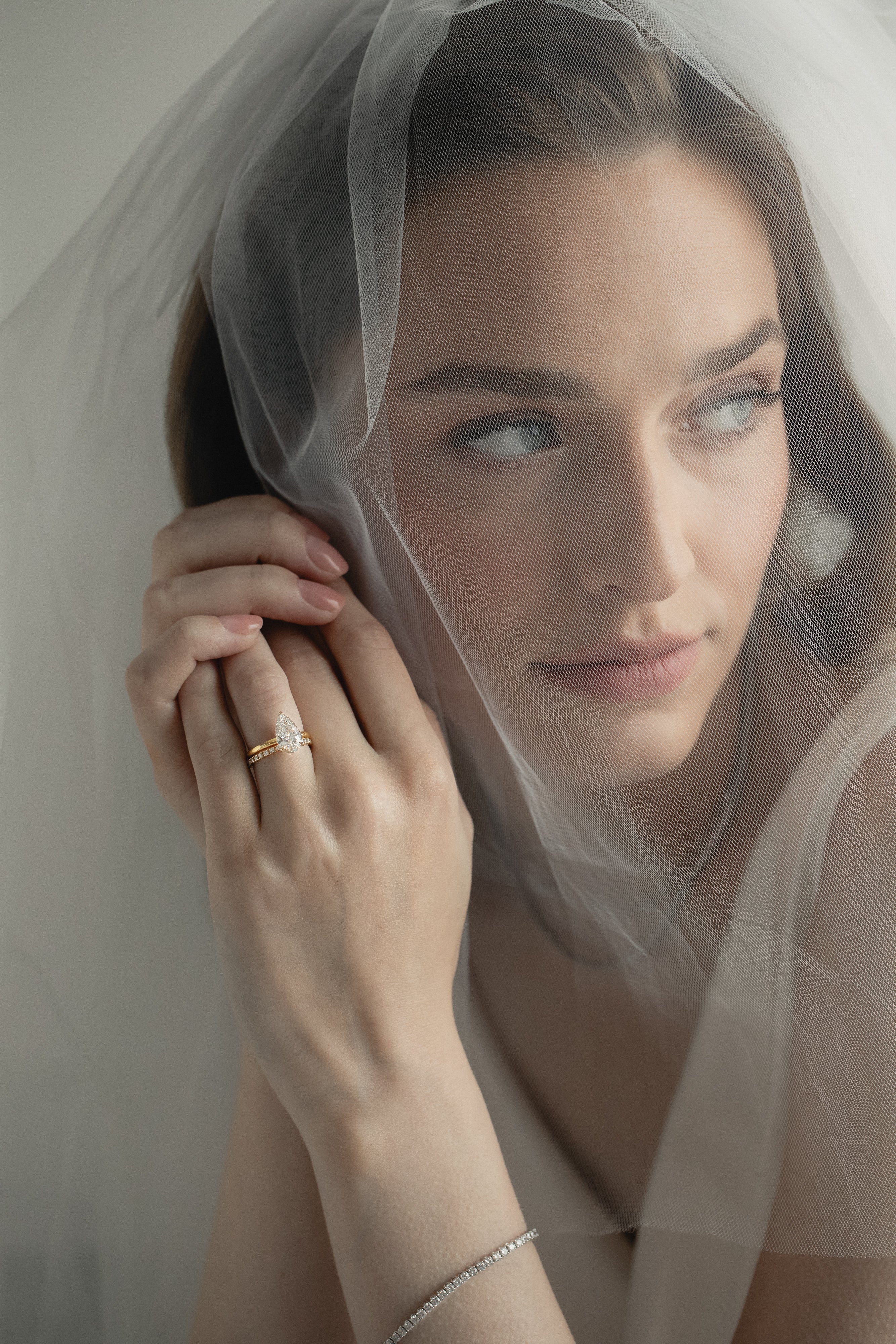 An editorial portrait of a bride looking through a sheer wedding veil, elegantly showcasing a KARAAT pear-shaped diamond solitaire engagement ring stacked with a matching rose gold wedding band.