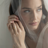 An editorial portrait of a bride looking through a sheer wedding veil, elegantly showcasing a KARAAT pear-shaped diamond solitaire engagement ring stacked with a matching yellow gold wedding band.