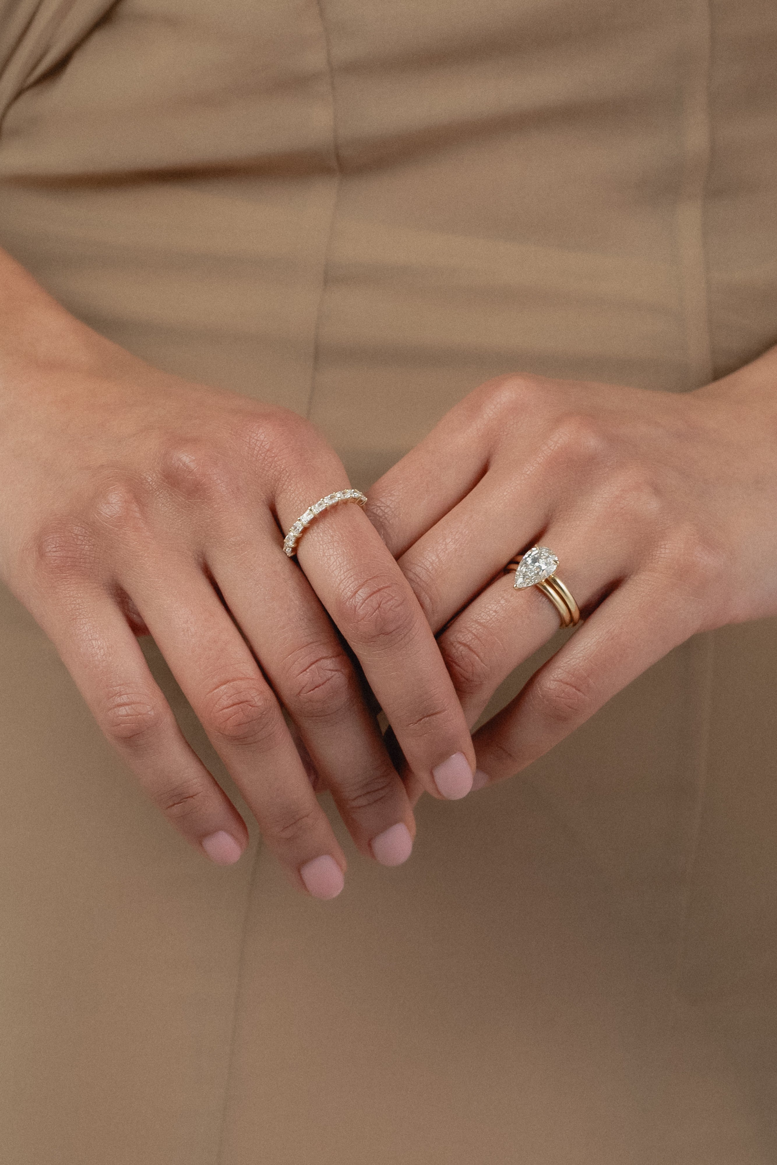 A close-up of a model's hands crossed against a beige dress, styled with a rose gold KARAAT pear-shaped diamond solitaire ring stacked with a gold band on one hand, and a radiant-cut diamond half eternity band on the other.