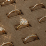 A close-up view of a KARAAT 18k yellow gold round solitaire diamond engagement ring with a pavé halo, beautifully displayed inside a plush beige velvet jewelry organizer tray alongside other wedding and engagement rings.