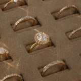 A close-up view of a KARAAT 18k rose gold round solitaire diamond engagement ring with a pavé halo, beautifully displayed inside a plush beige velvet jewelry organizer tray alongside other wedding and engagement rings.
