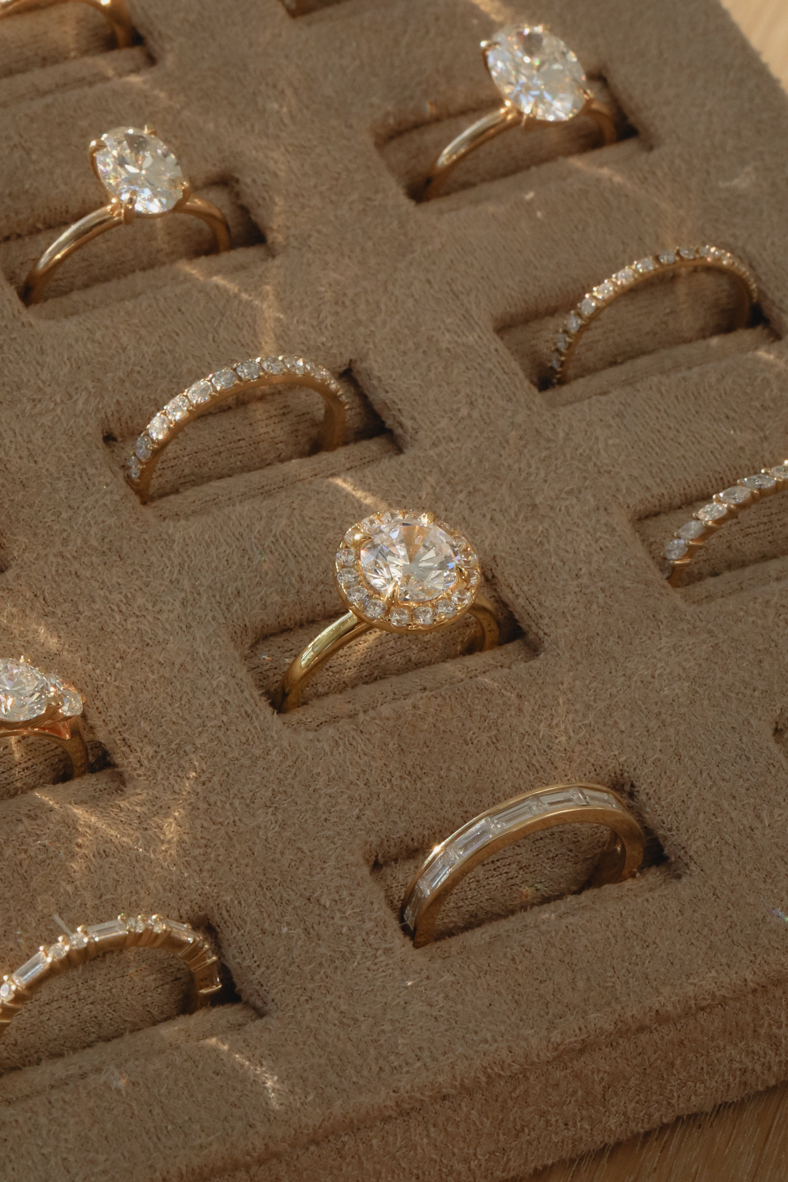 A close-up view of a KARAAT 18k rose gold round solitaire diamond engagement ring with a pavé halo, beautifully displayed inside a plush beige velvet jewelry organizer tray alongside other wedding and engagement rings.