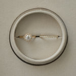 A flat-lay, top-down photograph of a white gold KARAAT round solitaire halo diamond engagement ring paired with a matching sparkling diamond wedding band, perfectly nestled side-by-side inside a round taupe velvet ring box.