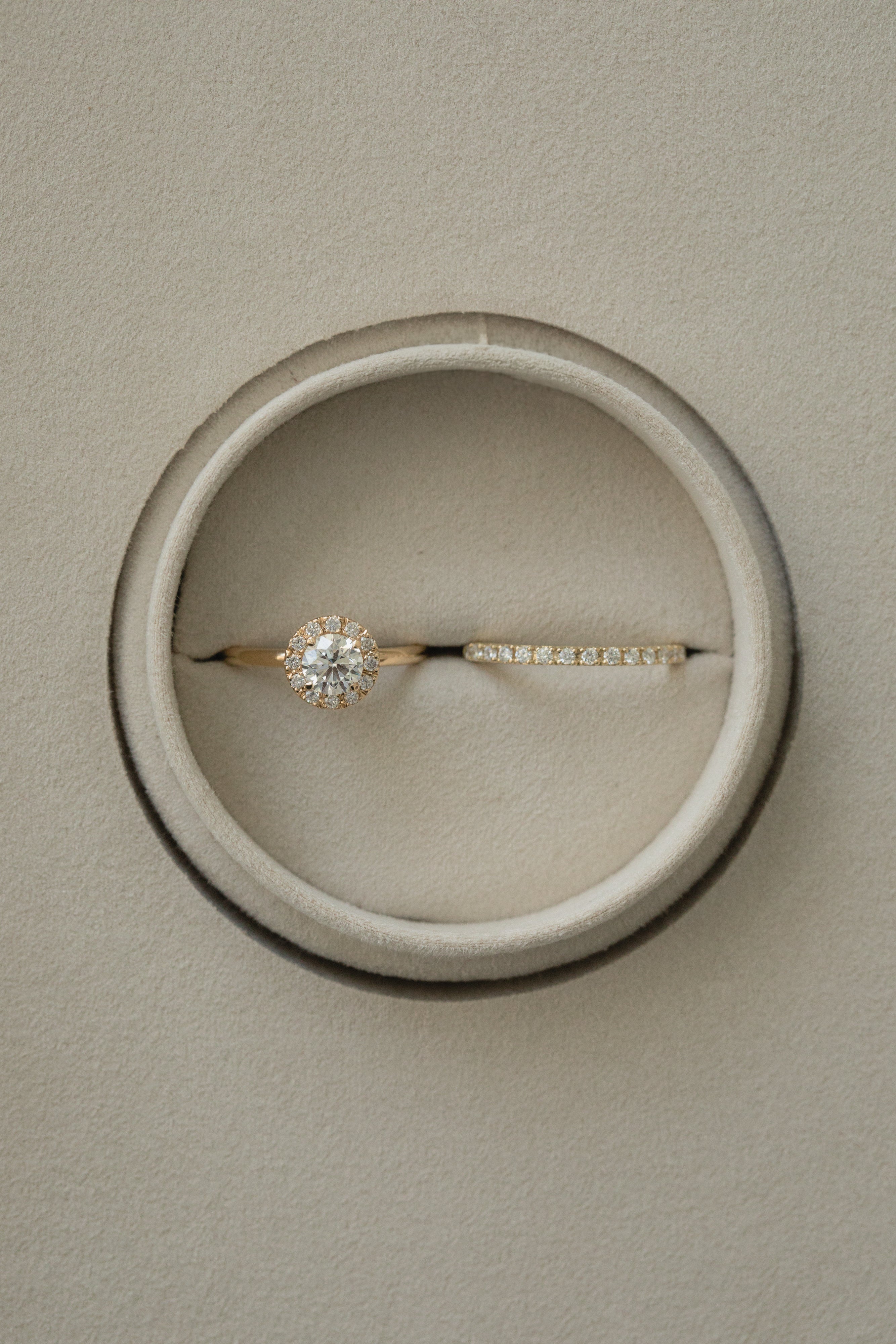 A flat-lay, top-down photograph of a white gold KARAAT round solitaire halo diamond engagement ring paired with a matching sparkling diamond wedding band, perfectly nestled side-by-side inside a round taupe velvet ring box.