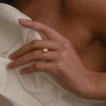 A close-up view of a model’s hand resting against a cream silk blouse, highlighting the elegant side profile, raised setting, and classic 18k gold band of the yellow gold KARAAT round solitaire halo diamond ring.