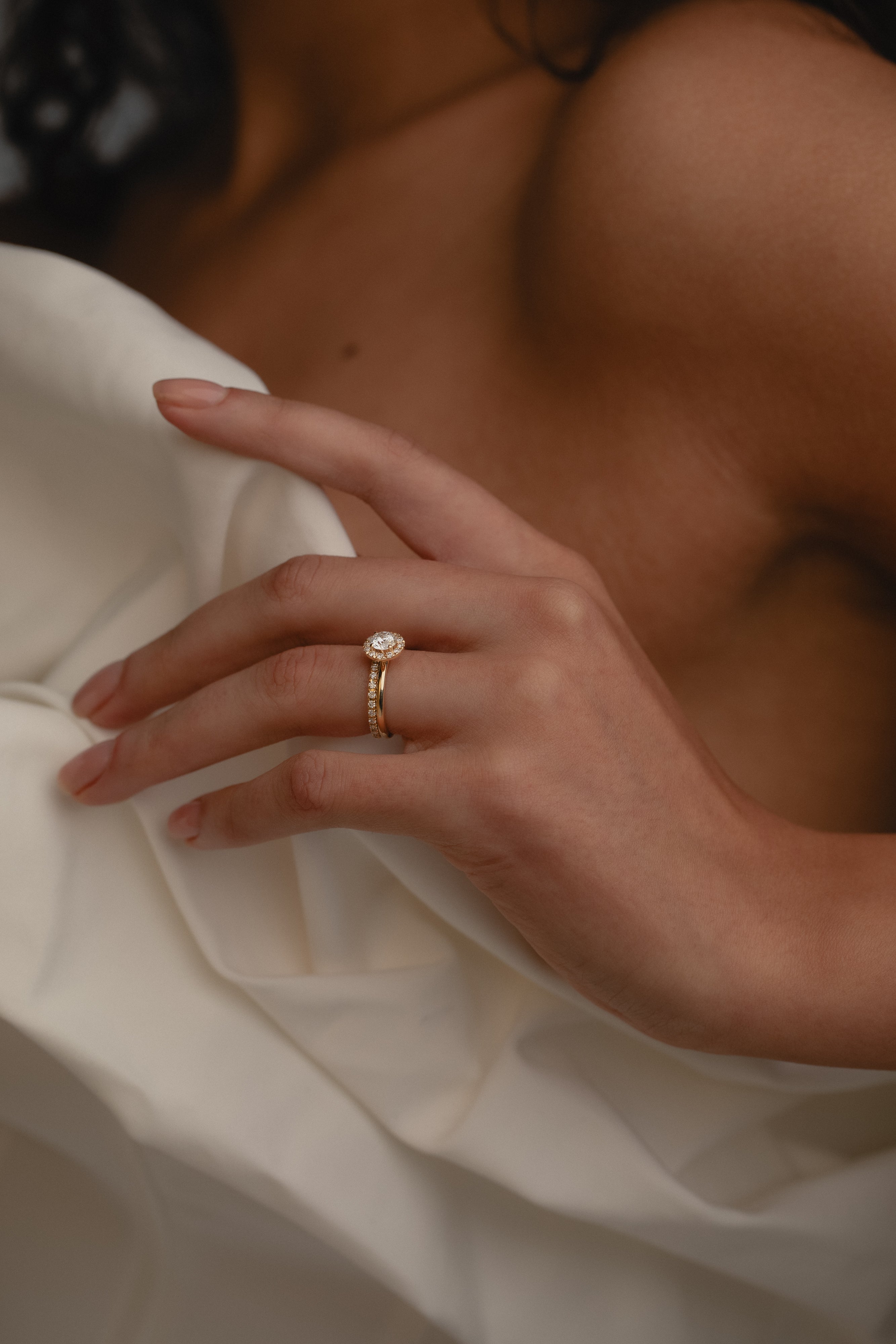 A close-up view of a model’s hand resting against a cream silk blouse, highlighting the elegant side profile, raised setting, and classic 18k gold band of the white gold KARAAT round solitaire halo diamond ring.