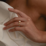 A close-up view of a model’s hand resting against a cream silk blouse, highlighting the elegant side profile, raised setting, and classic 18k gold band of the rose gold KARAAT round solitaire halo diamond ring.