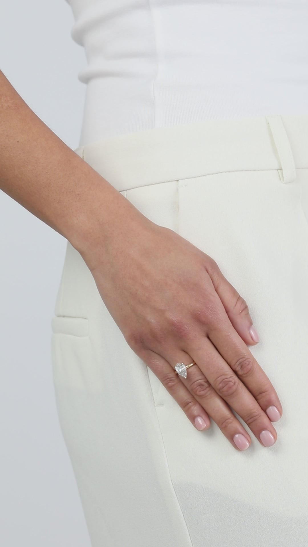 A clean product video showcasing a KARAAT pear-shaped diamond solitaire engagement ring on an 18k yellow gold band. The model has her hand casually placed in the pocket of tailored white trousers, capturing the brilliant sparkle and modern elegance of the ring against the crisp white fabric.