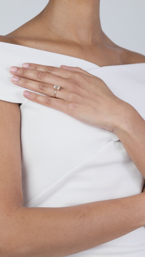 A elegant product video showcasing a KARAAT emerald-cut solitaire diamond engagement ring on an 18k white gold band. The model wears a white off-the-shoulder top and rests her hand on her chest, beautifully capturing the clean lines and flash of the diamond.